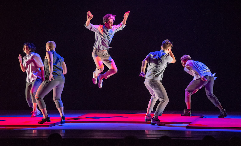 A dancer leaps in the air with arms near his head. Other dancers in cropped beige pants and button up shirts tap near him. Lights cast a hot pink glow on floor they dance upon.